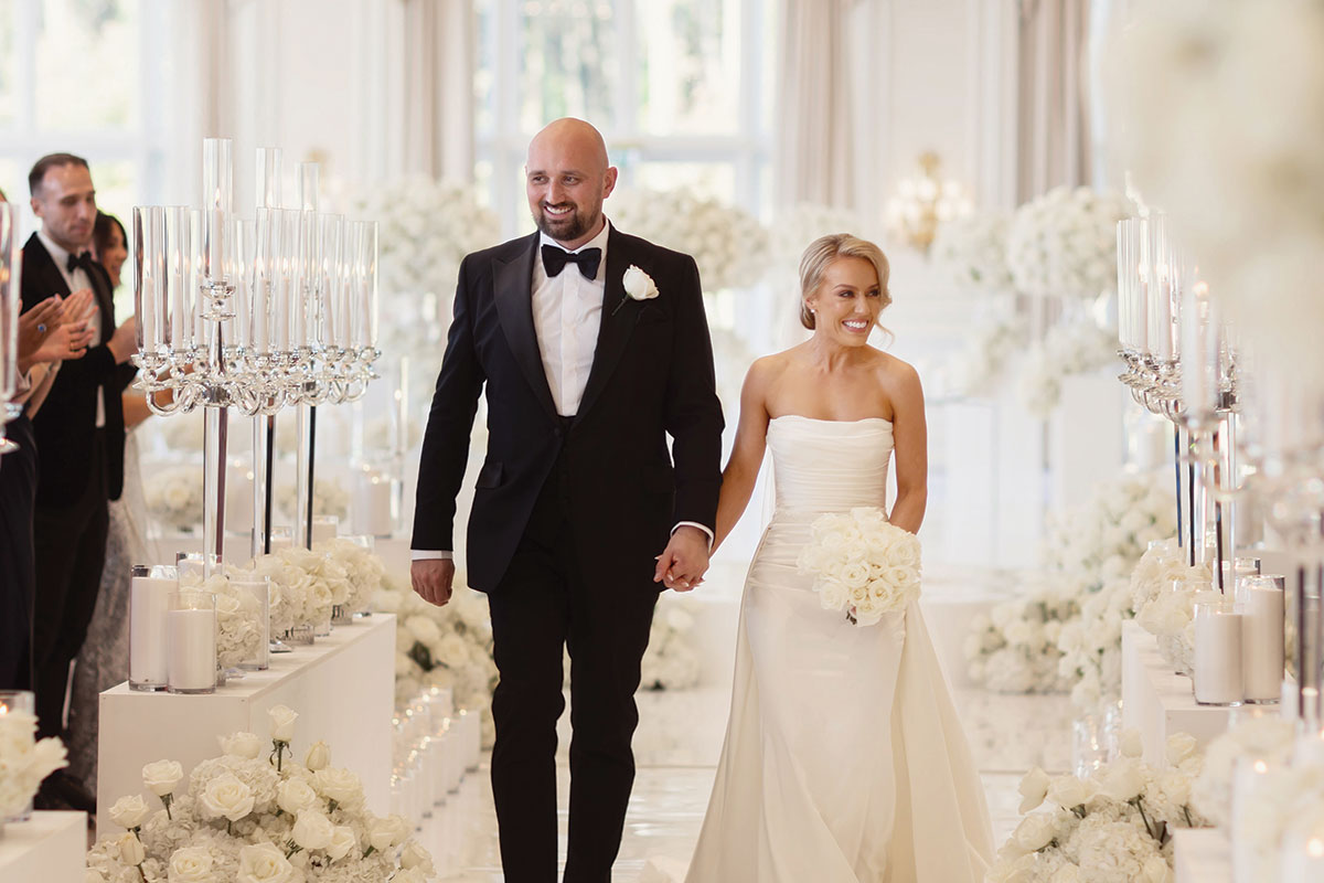 a bride wearing a strapless wedding dress and veil holding hands with a groom wearing a black tuxedo suit in a room filled with white flowers and candles in glass hurricane vases after their wedding ceremony at Trump Turnberry