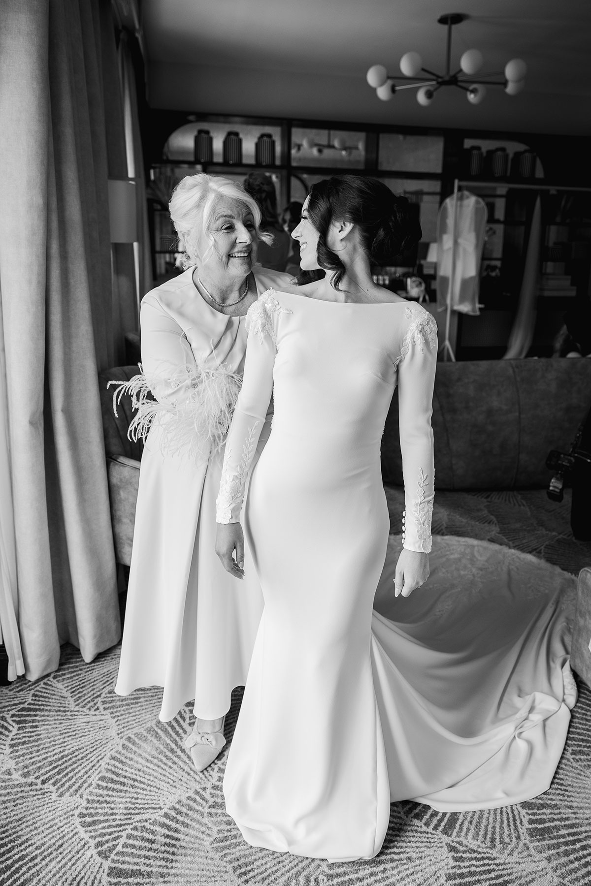 Black-and-white photo of the bride smiling as her mother helps her into a long-sleeved wedding dress