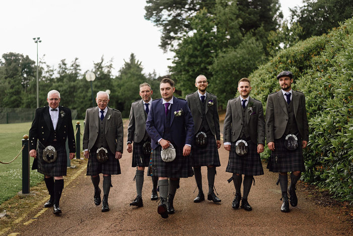 groom and six groomsmen walk in a side by side in a line all wearing full tartan outfits