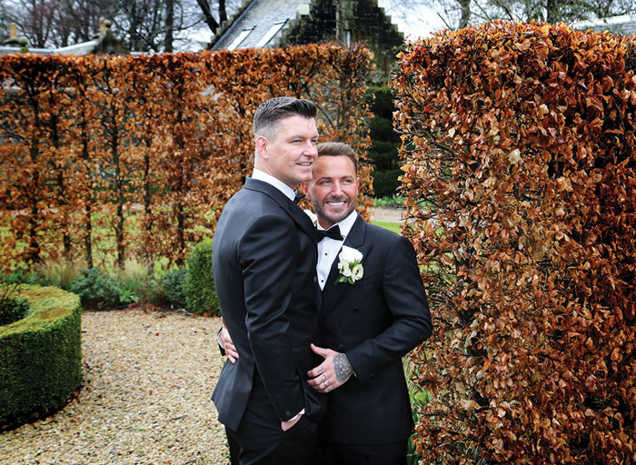 two grooms wearing smart black suits and bow tie embrace. There are tall hedges with russet leaves in the background and gravel on the ground