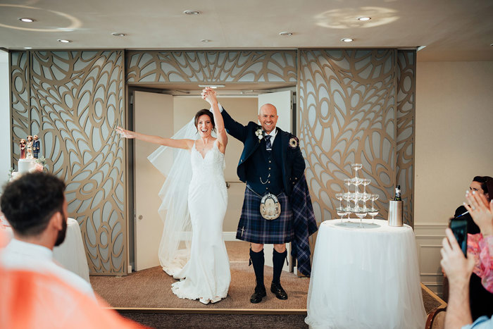 a cheering bride and groom with hands in air walk through a door on an ornate wood-panelled background at seamill hydro