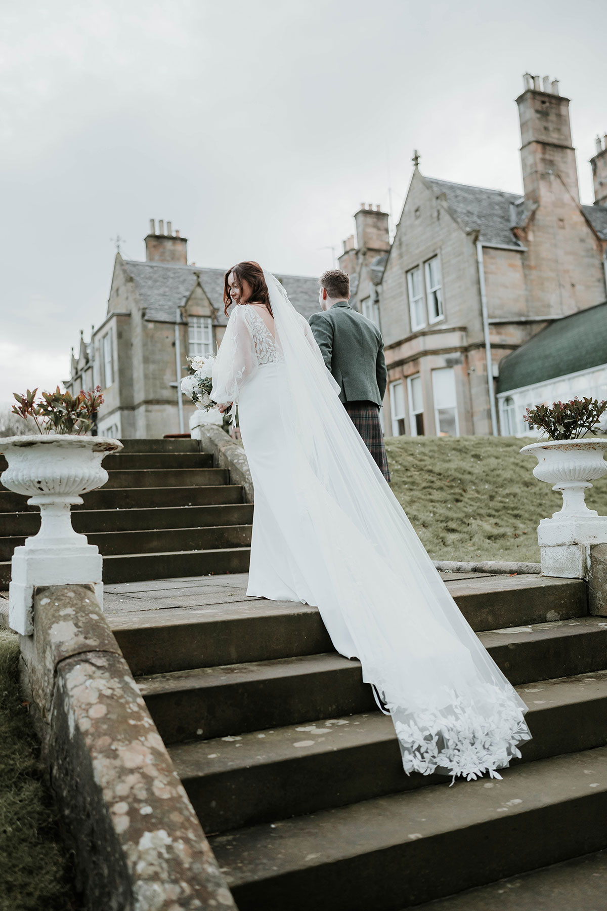 Bride’s long veil draped across the stone steps outside Norton House as she walks with the groom.