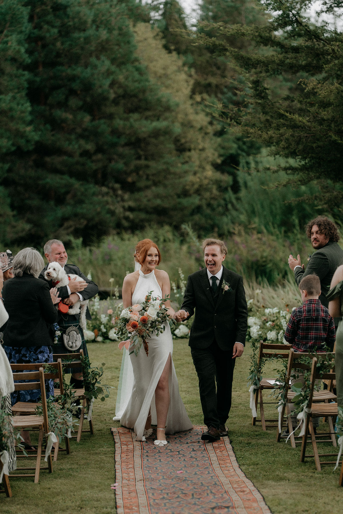 Bride and groom walking back down outdoor ceremony aisle as guests applaud