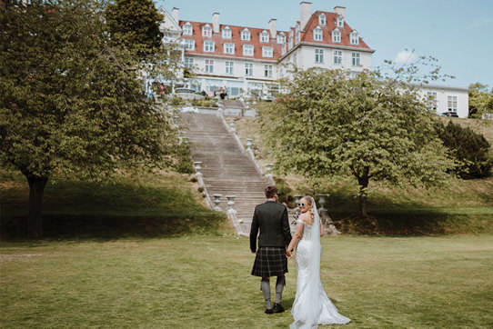 A groom in a kilt and a bride in a white dress stand on a grass lawn at the bottom of a tall set of stone stairs leading up to a large white building with an orange roof