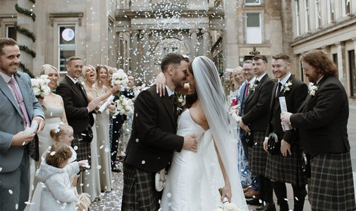 A bride and groom standing outside surrounded by their wedding guests as white confetti is thrown over them