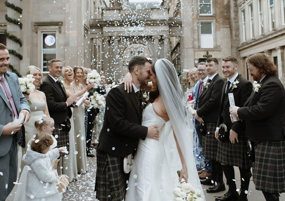 A bride and groom standing outside surrounded by their wedding guests as white confetti is thrown over them