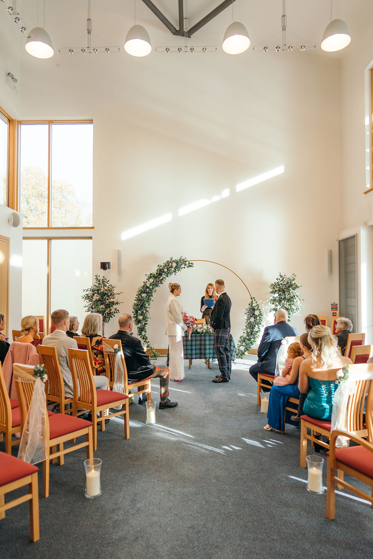 Couple stand at modern indoor Scottish wedding ceremony with circular floral arch and seated guests