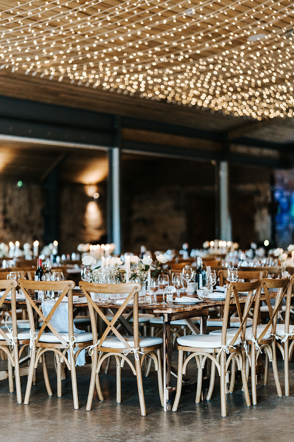 Rustic wedding reception tables with wooden chairs, candlelight and overhead fairy-light canopy