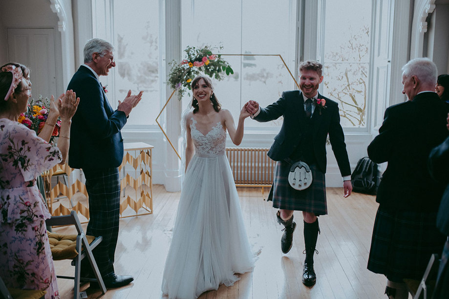 a jubilant bride and groom walking up the aisle after wedding ceremony at Netherbyres House as guests clap. There is a hexagonal frame with flower decoration behind them.