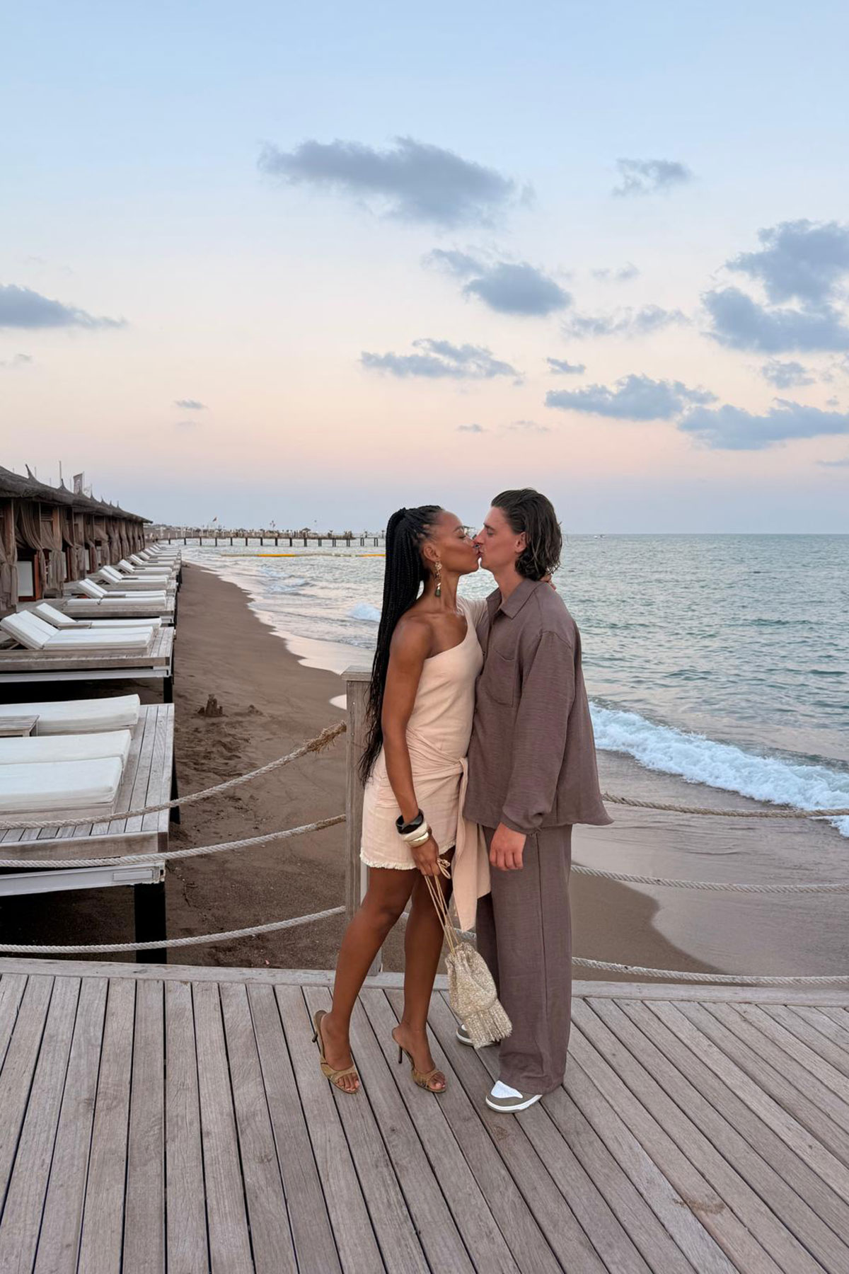 A couple share a kiss on a wooden deck beside a sandy beach at sunset, with cabanas and the sea in the background