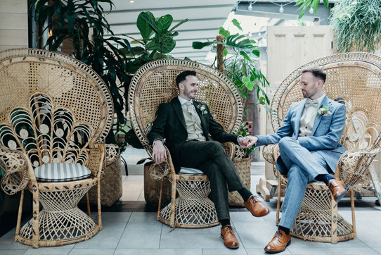 two groom sitting on wicker peacock chairs holding hands