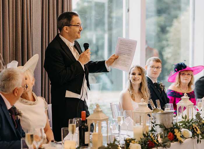 A man holding a piece of paper and a microphone making a speech at a long table of other people