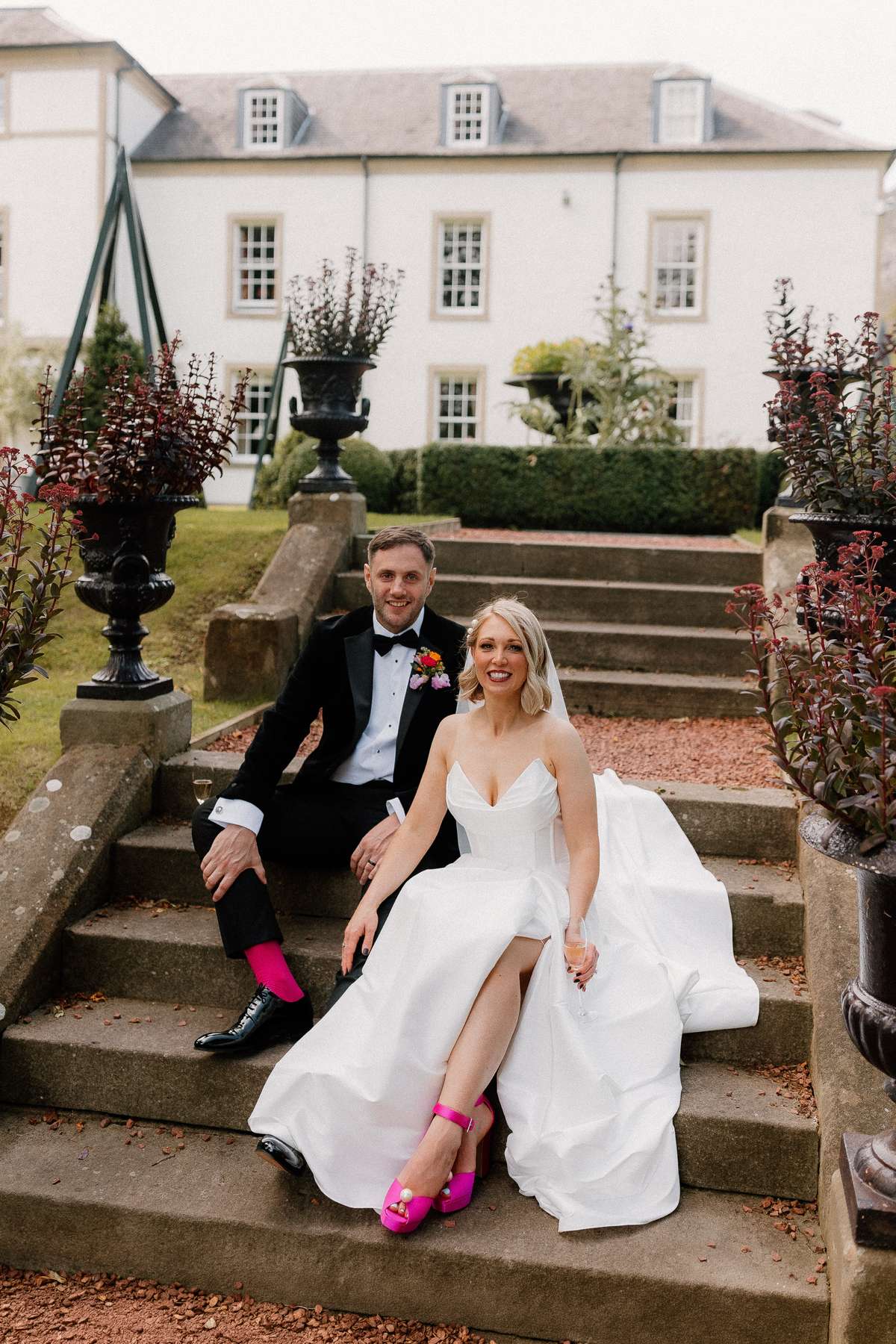 Newlyweds Kara and Rory sit on the garden steps at Prestonfield House in Edinburgh, with Kara wearing bright pink wedding shoes.