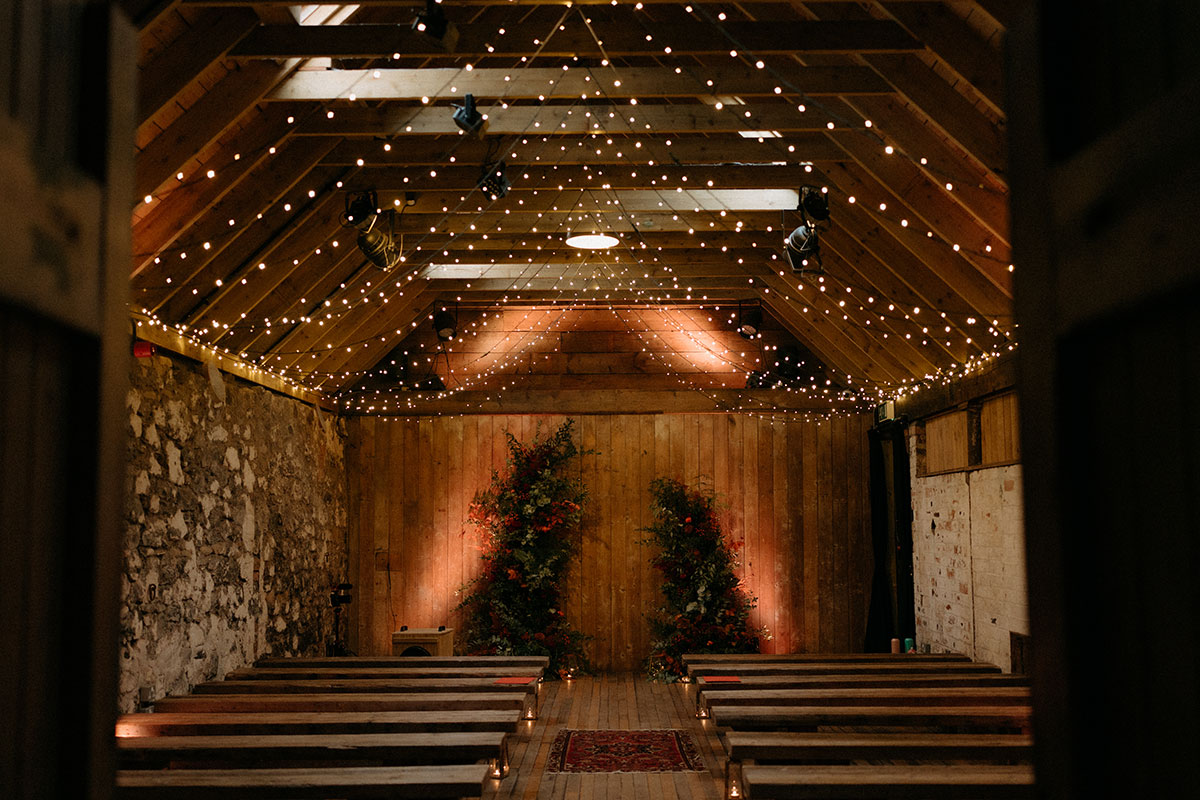 A wooden-floored barn with stone walls, fairy lights and bench seating arranged for a ceremony.