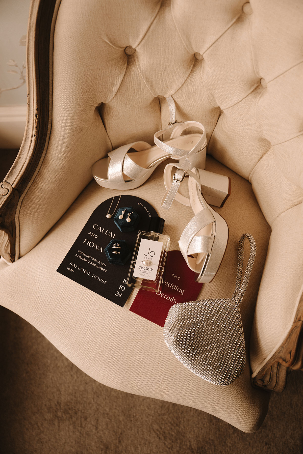 Bridal flatlay with wedding shoes, jewellery, Jo Loves perfume, silver bag, and wedding invitations on chair.