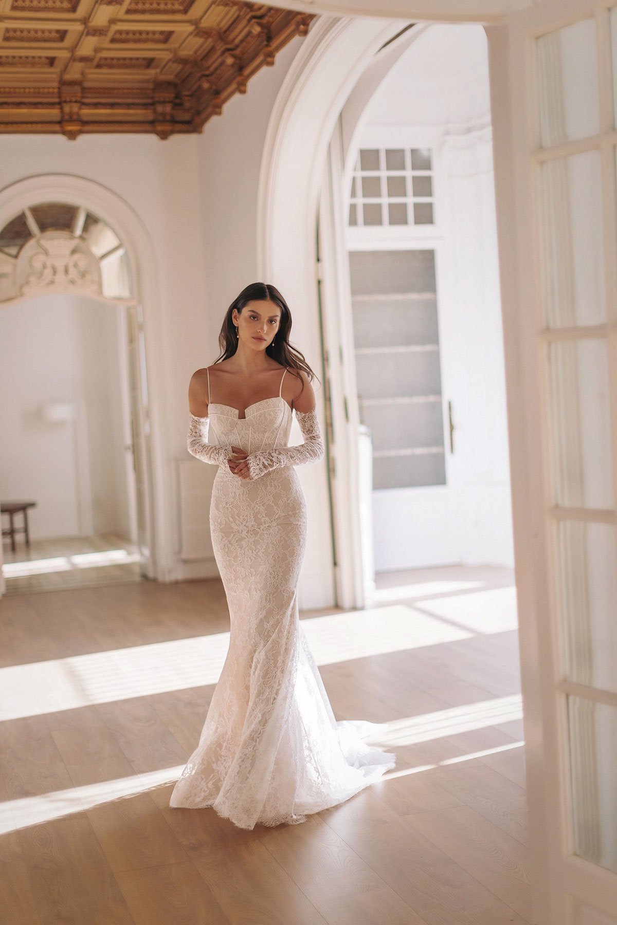 Bride wearing a fitted lace wedding dress with off-the-shoulder sleeves by Opus Couture in a bright interior setting.