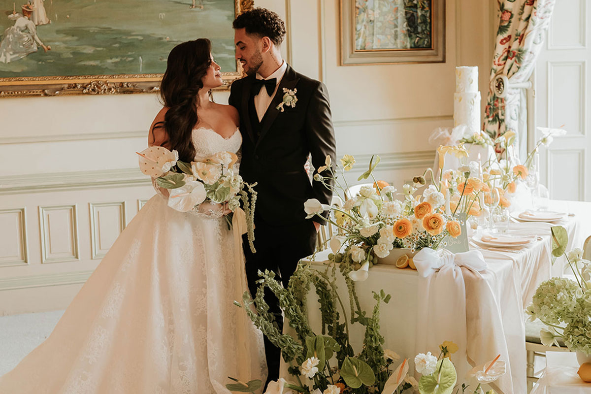 bride in a strapless long lace wedding dress stands next to groom in black tux and a large table filled with green, orange and white florals, white bows and a two tier white wedding cake in the background