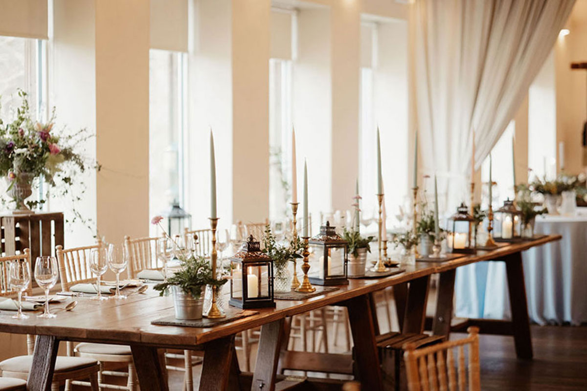 long wooden table decorated with candlesticks, lanterns and plant pot flowers alongside glassware and cutlery