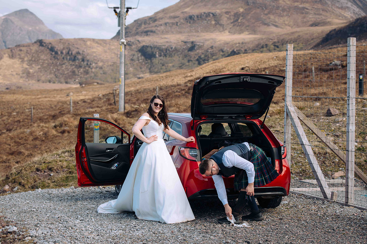 Bride in wedding dress and groom in kilt change shoes beside red car in the rugged Highlands after beach ceremony.