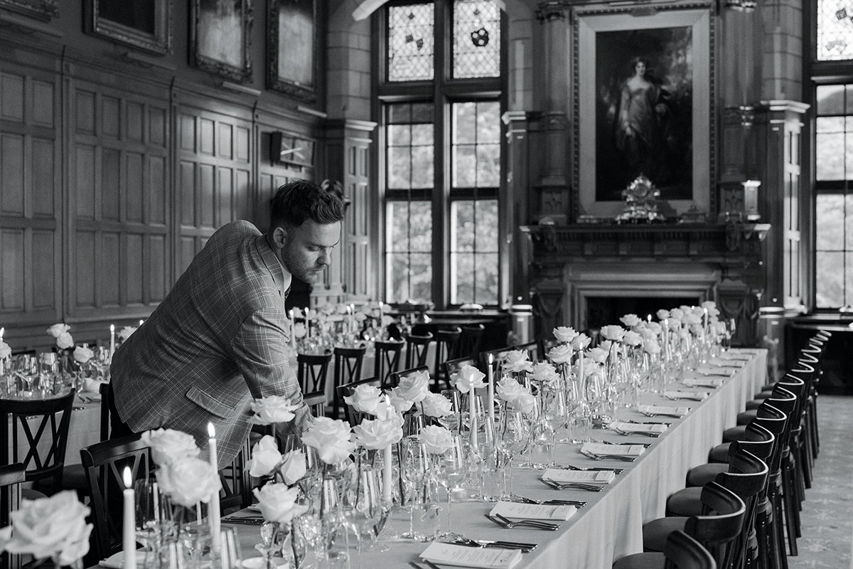 Wedding planner adjusting long dining tables styled with white roses, candles and place settings inside an elegant historic dining hall