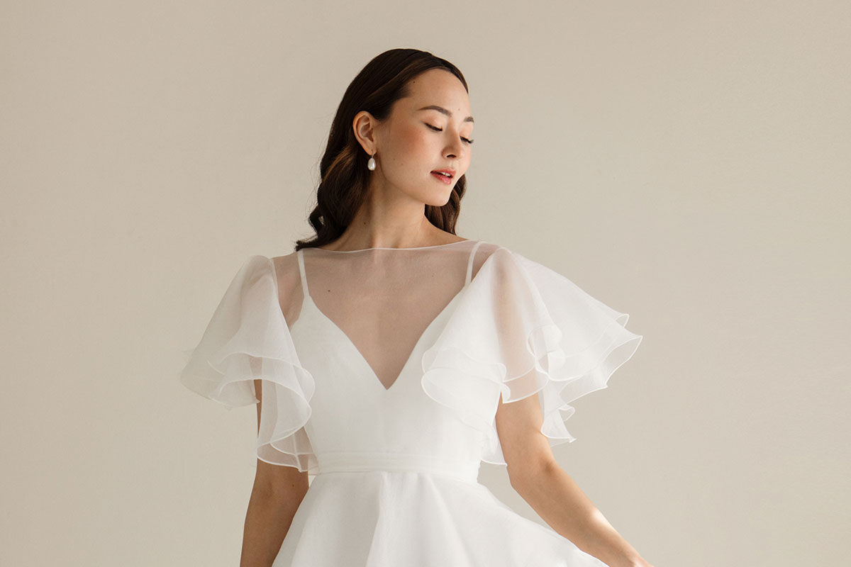 dark haired model wears v-neck white wedding dress with sheer layer over bodice providing fluttery sleeves