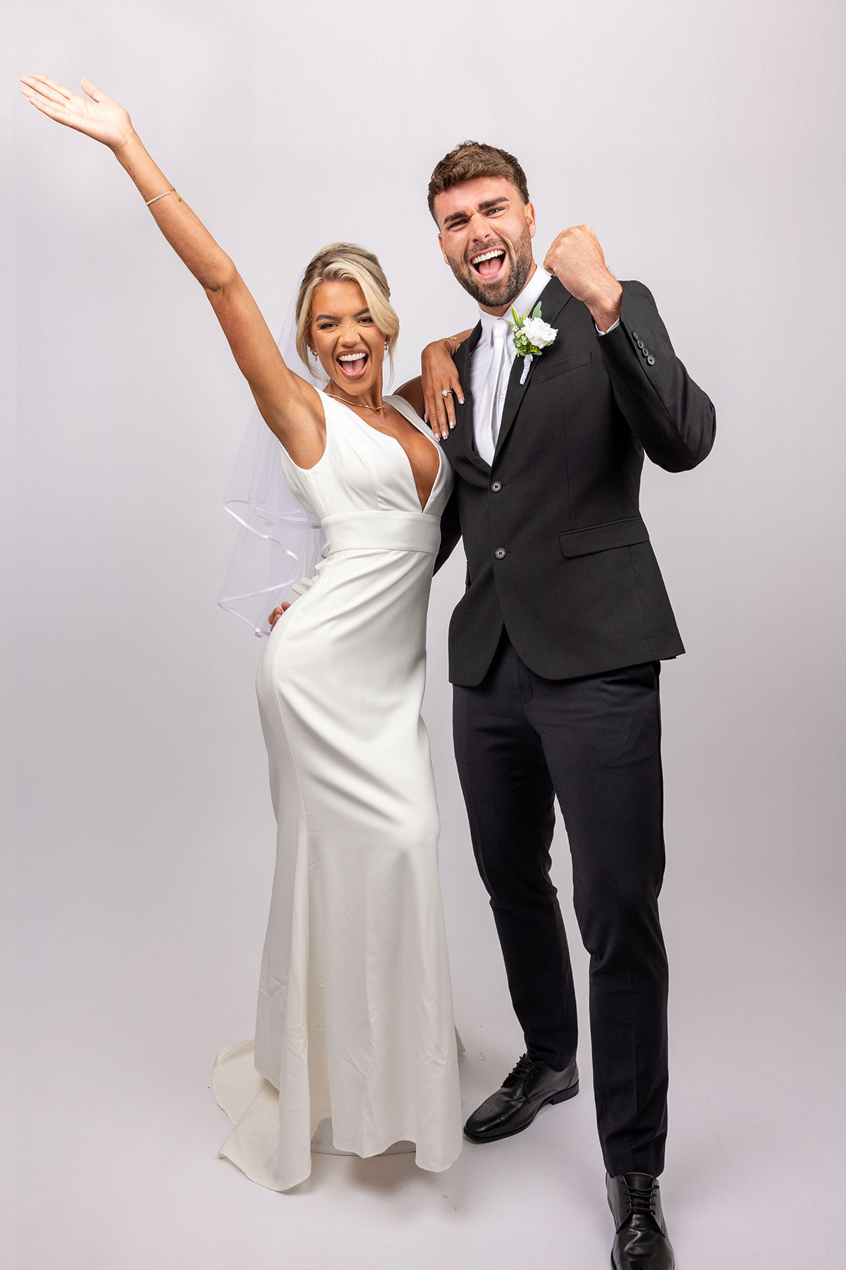 Bride and groom pose together in a studio, celebrating with raised arms and joyful expressions; the bride wears a sleek white gown and veil while the groom is in a classic black suit with boutonnière