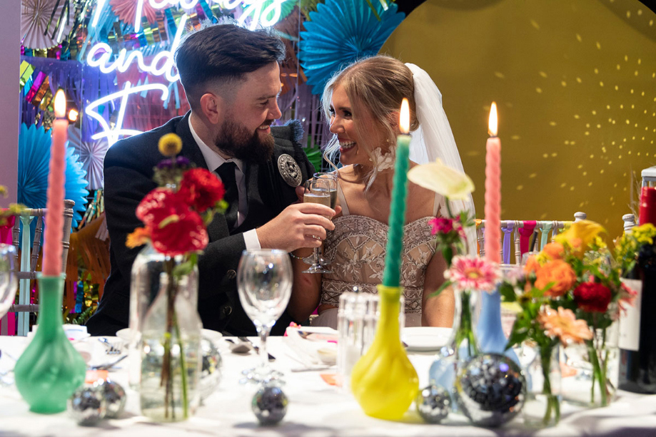 Bride and groom smile at each other at top table which is covered with colourful candles