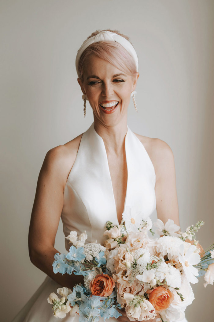 a smiling bride wearing a smooth white halter neck dress, holding a large pastel blue and peach and white bridal bouquet.