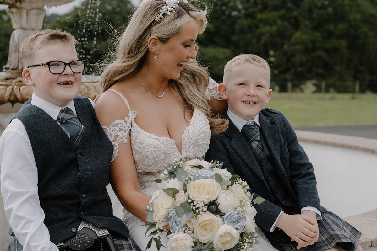 bride holding a white and blue bouquet looks towards younger boy next to water fountain at cornhill castle
