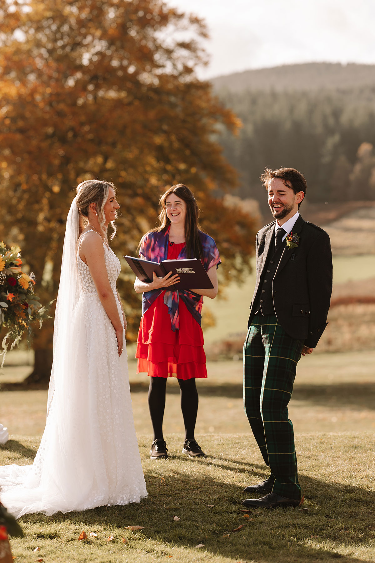 Bride and groom smiling with celebrant during wedding ceremony in autumn countryside.