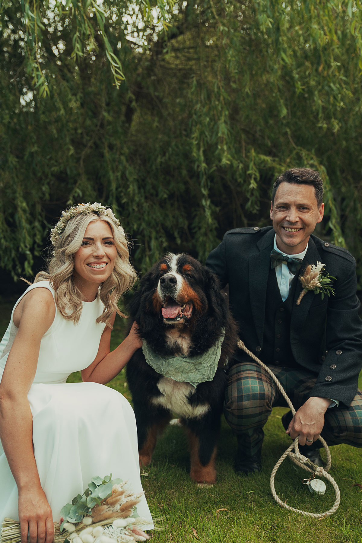 Bride and groom crouch beside their large Bernese mountain dog, all smiling at the camera, with greenery in the background