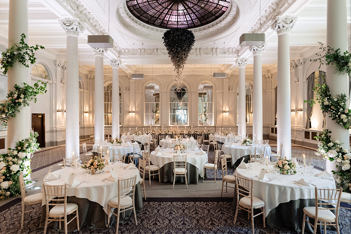 a large space with high ceilings, a dome centre and eight classic white columns is set with formal round tables and a variety of white flowers and greenery throughout