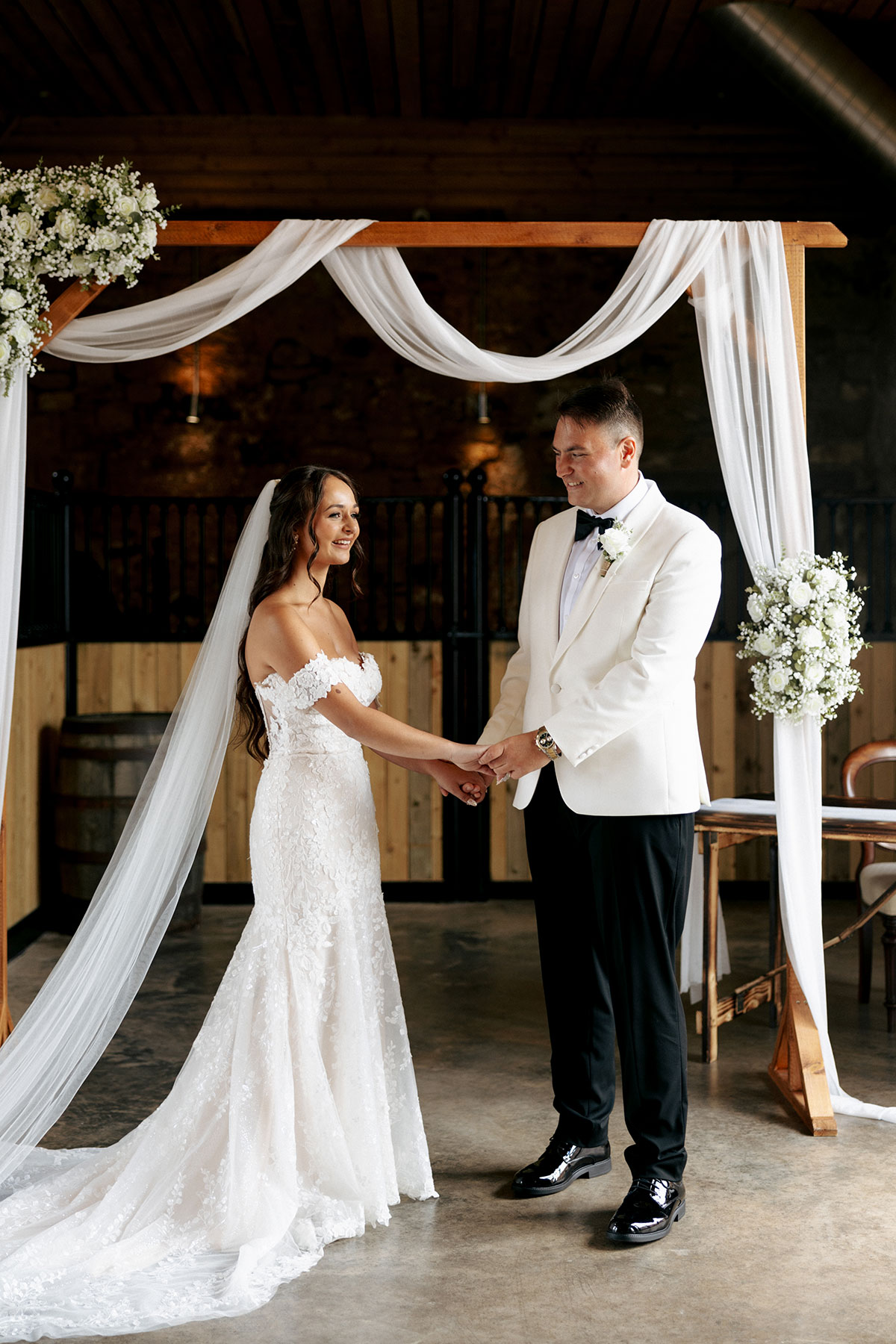 Bride and groom exchange vows beneath a wooden arch draped with white fabric and flowers at Falside Mill.