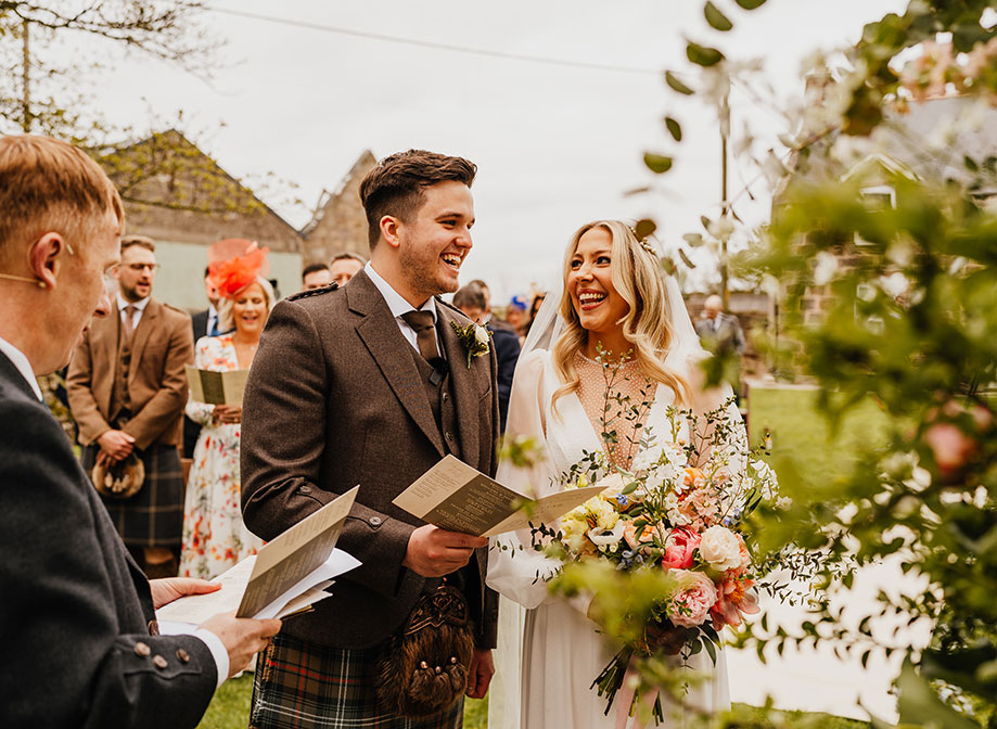 a bride and groom hold printed card orders of ceremony and look happily at one another during their outdoor wedding ceremony. Wedding guests stand in the background