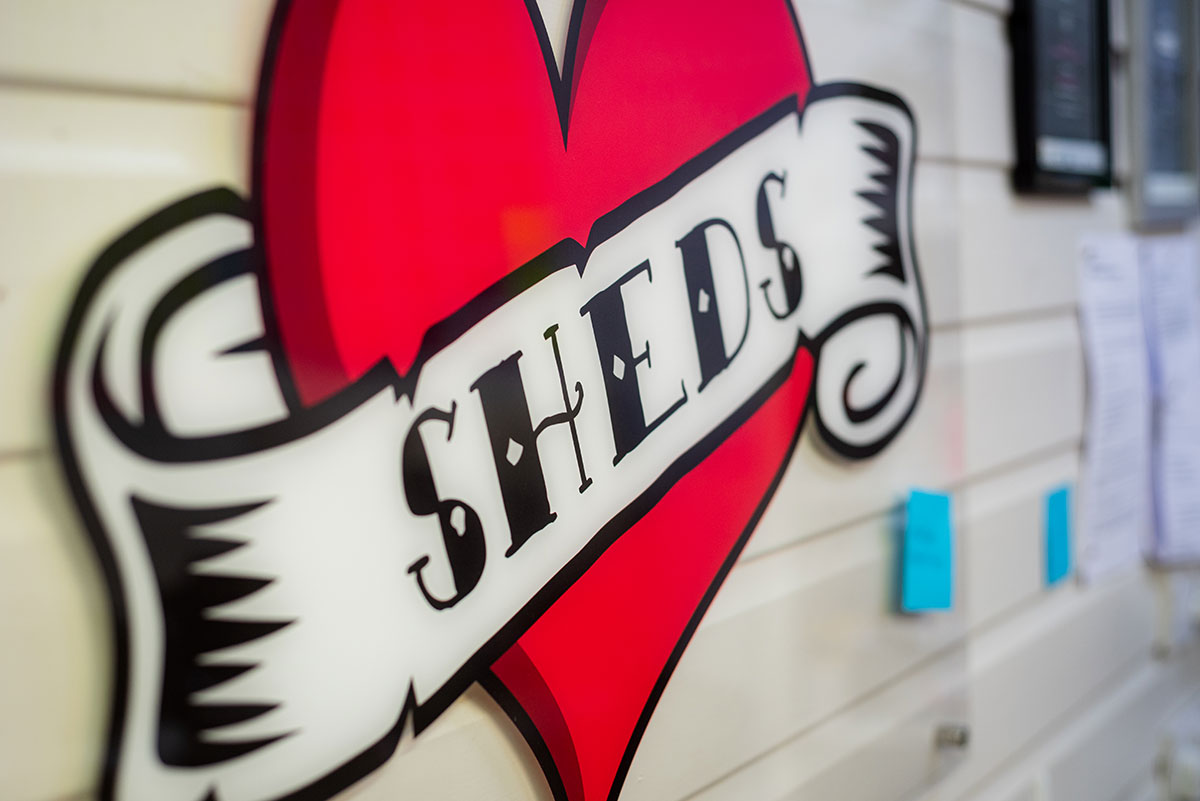 Gillies & Mackay Red Heart Sign Reading SHEDS Hanging On An Ivory Cladding Wall