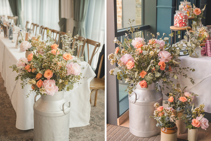 Orange and pink flowers sit in milk pails and flower pots sit together in front of tables with white table cloths