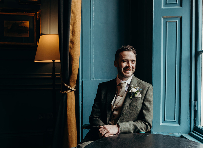 a laughing groom wearing a green tweed suit poses by a teal-painted window and rust-coloured curtain