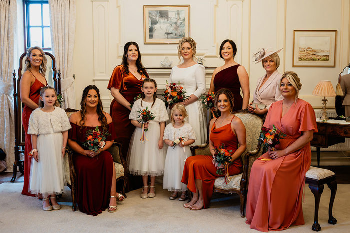 bride in white wedding dress stands in the centre of formal photo set up joined by young flower girls in white, bridesmaids in red dresses, and a mother of the bride in a pink outfit and matching hat