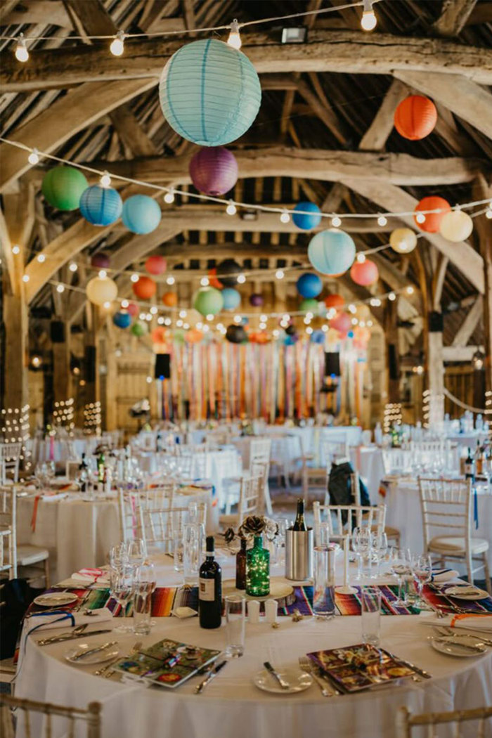 a barn with large wooden rafters with colourful paper larners and festoon lights hanging from them, with round tables with white tablecloths on the floor below