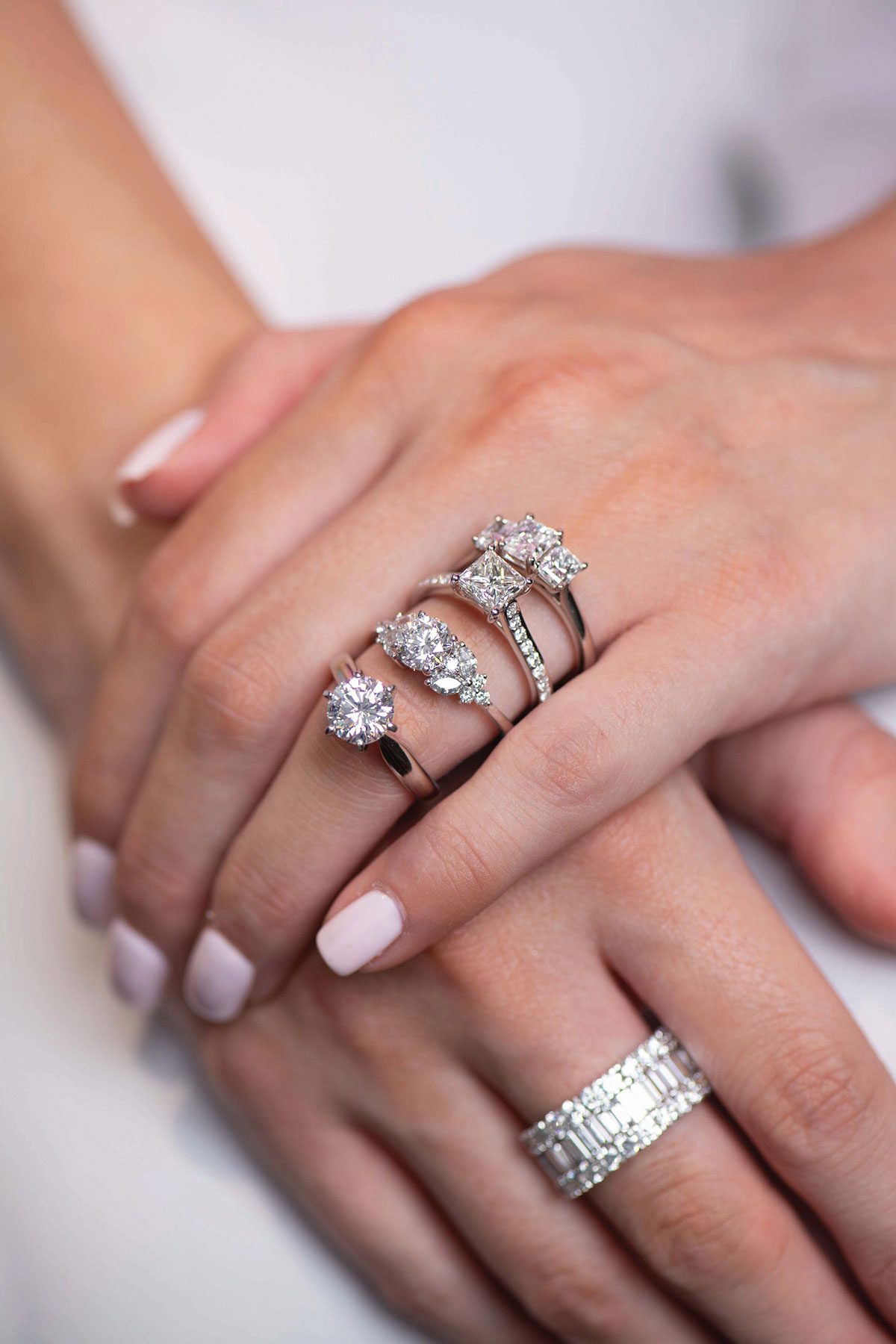 close up of two hands laid across each other, the left hand ring finger modelling four diamond rings and the right hand a diamond wedding band
