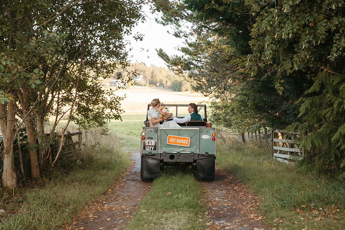 Newlyweds drive away in vintage Land Rover with Just Married sign after Highland farmhouse wedding in Nethy Bridge