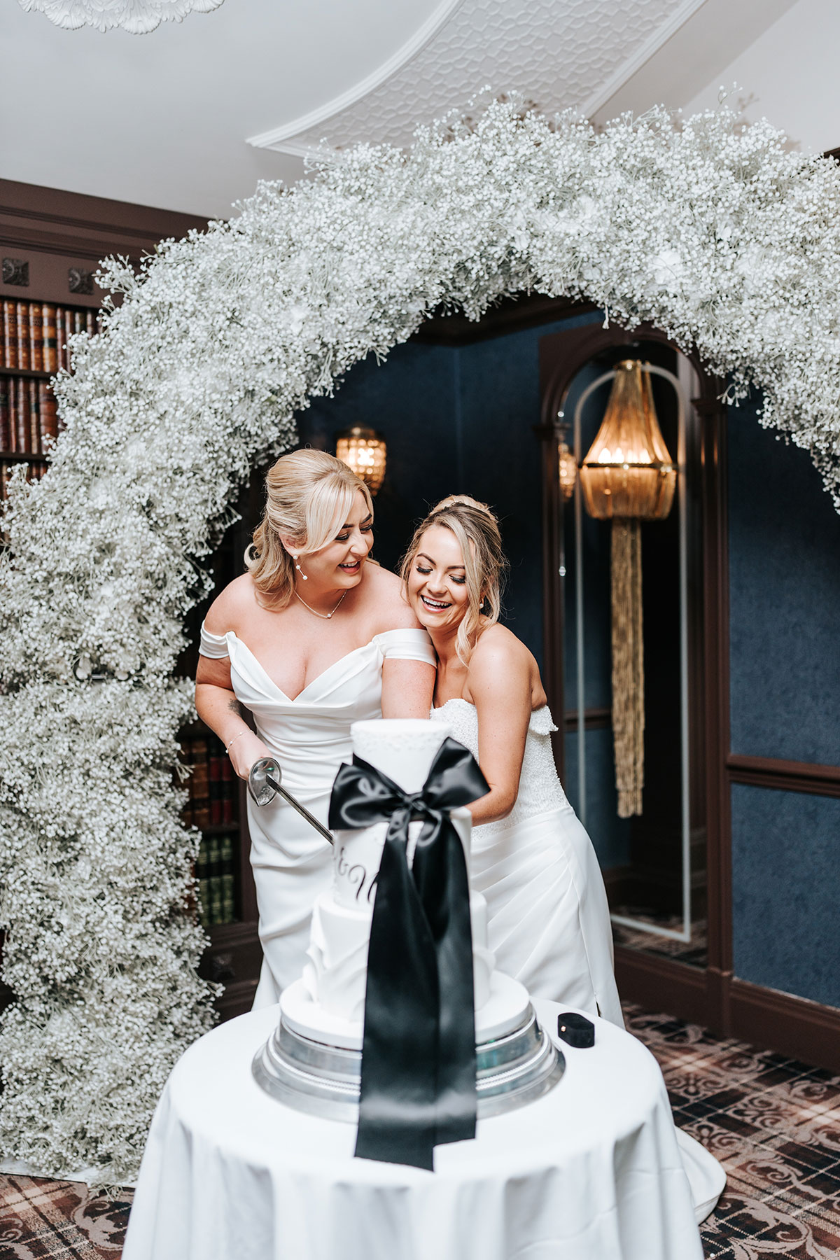 Newlywed brides cutting their wedding cake beneath a dramatic white floral arch at their reception