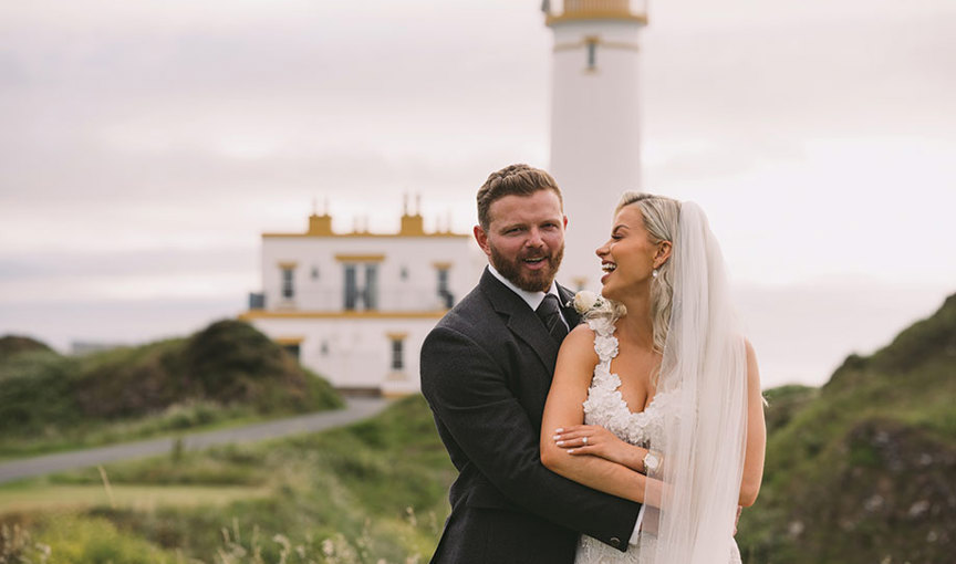 A laughing bride wearing an ivory dress and veil, and groom in dark grey kilt outfit embrace in front of Turnberry Lighthouse