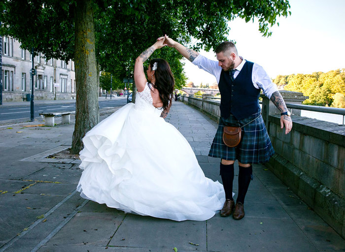 A woman in a wedding dress with a large skirt being spun around by a man in a blue and green kilt 