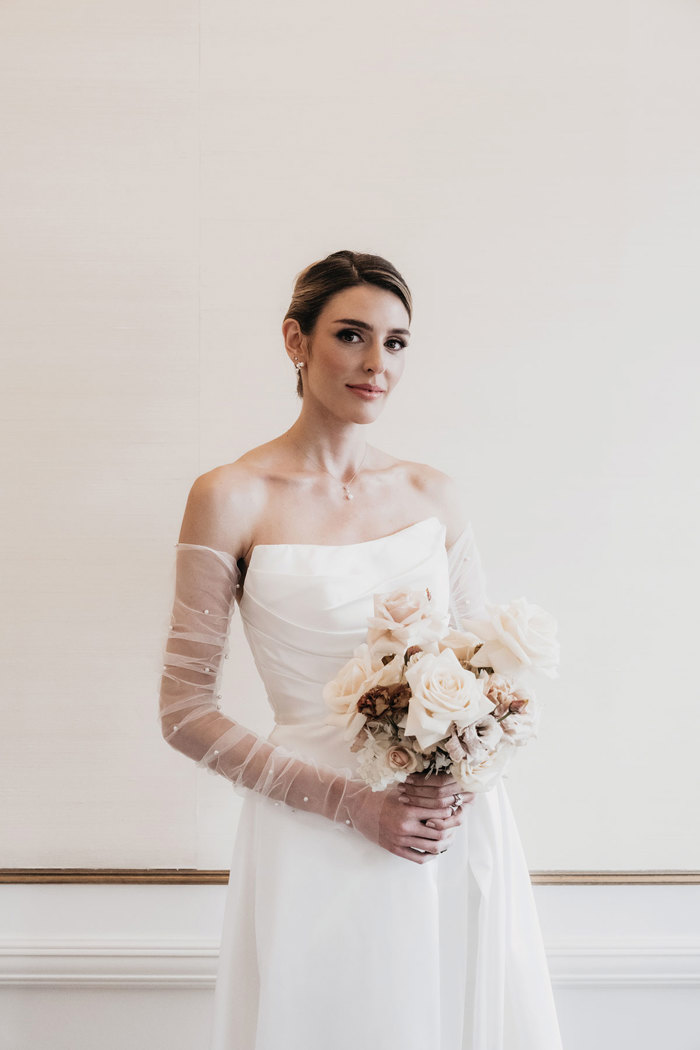 a brunette bride wearing a strapless ruched bodice wedding dress with removable tulle lace sleeves from Kudos Bridal Dunfermline. She is standing in front of a neutral coloured wall.