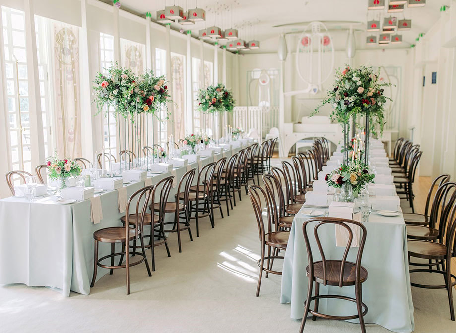 an elegant pastel room designed by Charles Rennie Mackintosh set for a wedding dinner. There are two long tables flanked by dark wooden chairs and tall pink and green floral arrangements placed on each table.