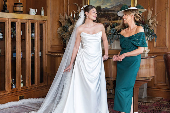 A bride wearing a strapless white wedding dress and long veil and a mother of the bride wearing forest green dress