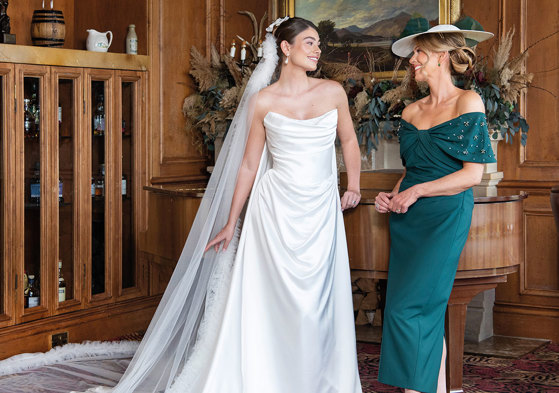 A bride wearing a strapless white wedding dress and long veil and a mother of the bride wearing forest green dress