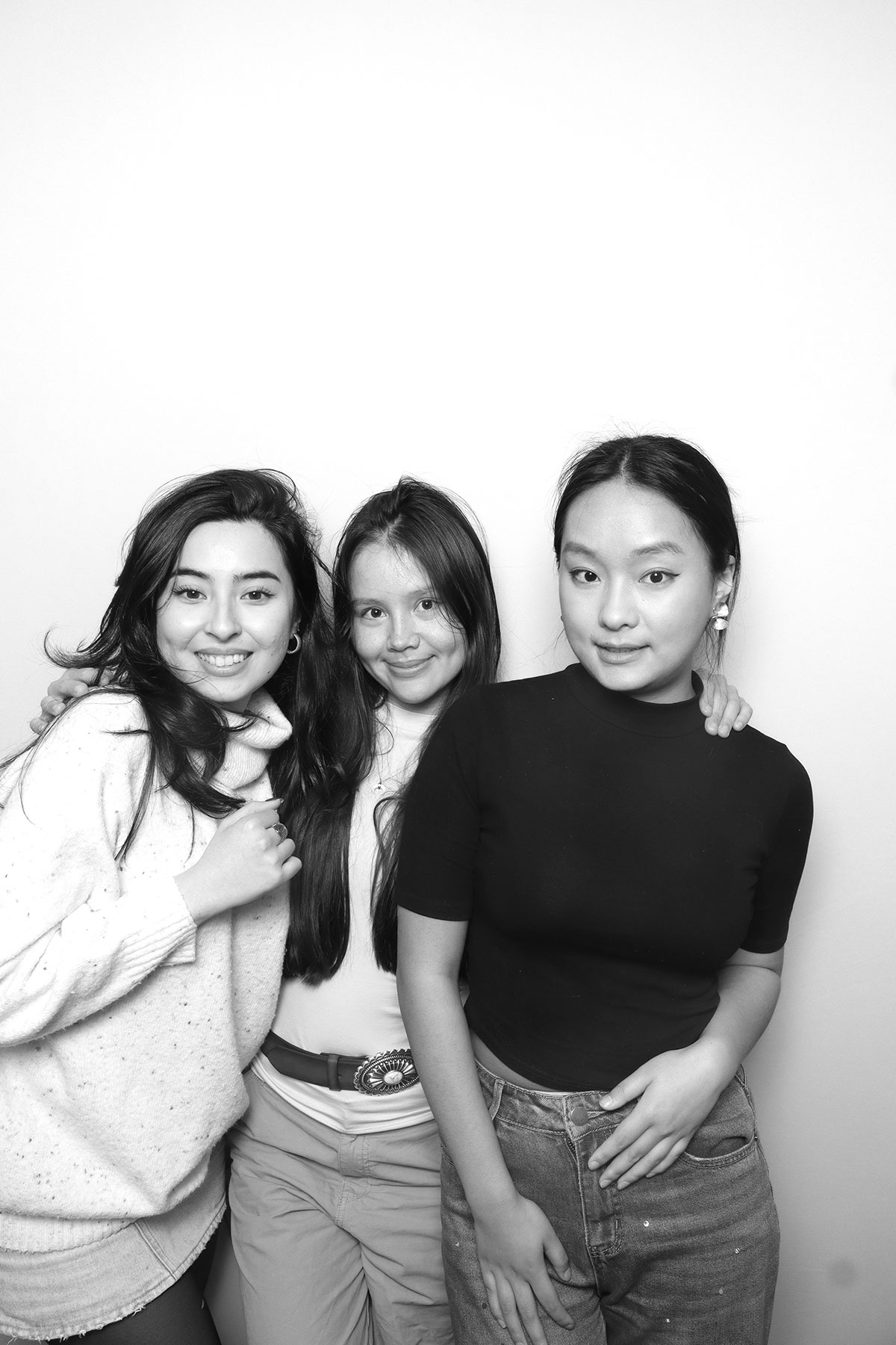 Black and white photo booth portrait of three women smiling together against plain backdrop