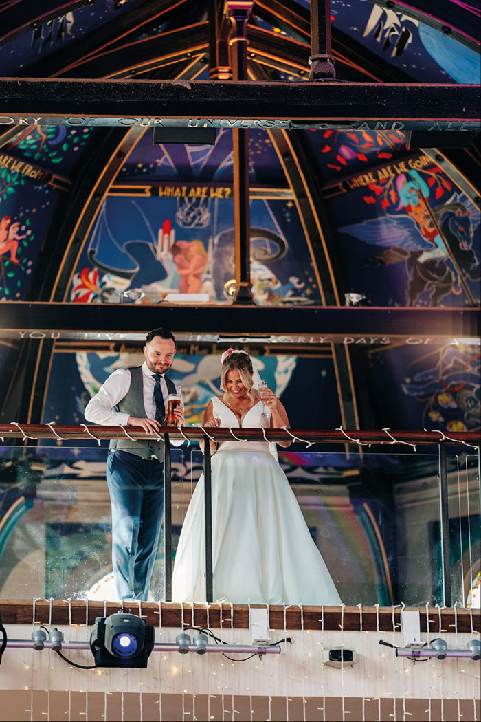 bride and groom couple each holding a drink in their hand, stand on a balcony, with large colourful ceiling above them, as they look down beyond the clear railing